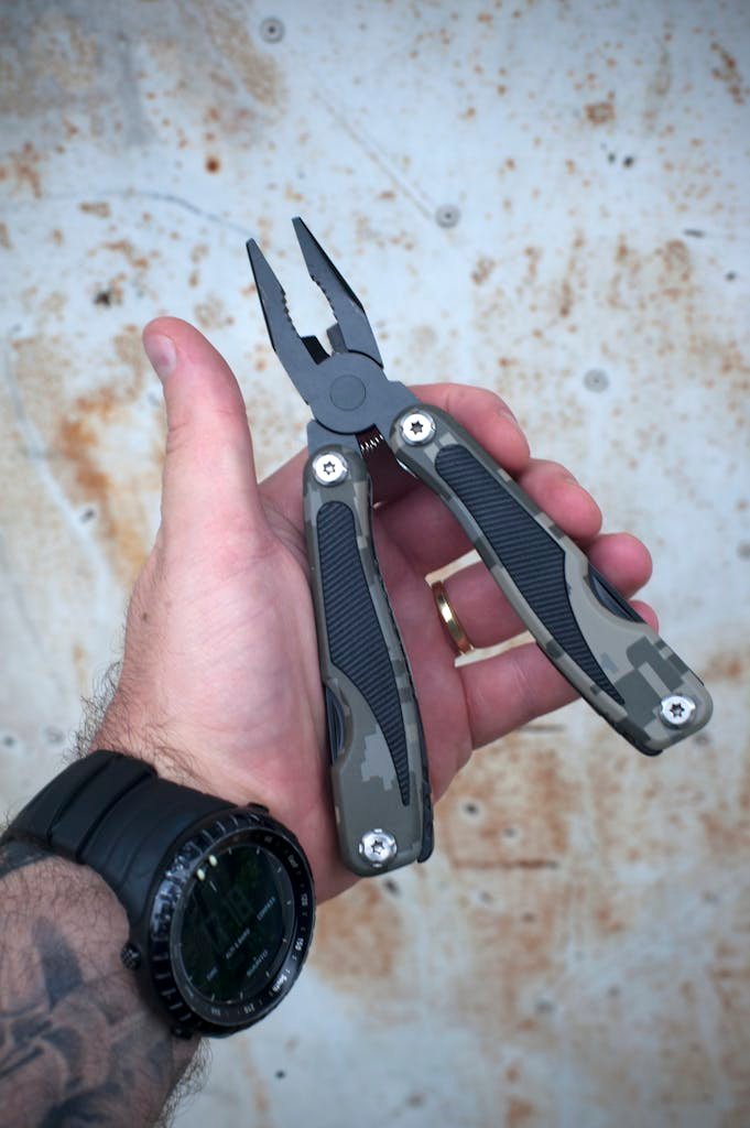 Photo by Dan Galvani Sommavilla A close-up shot of a hand holding a camouflaged multi-tool pliers against a rusted metal surface.