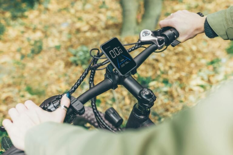 Person riding a bicycle in a forest with autumn leaves, close-up of handlebars and GPS device.